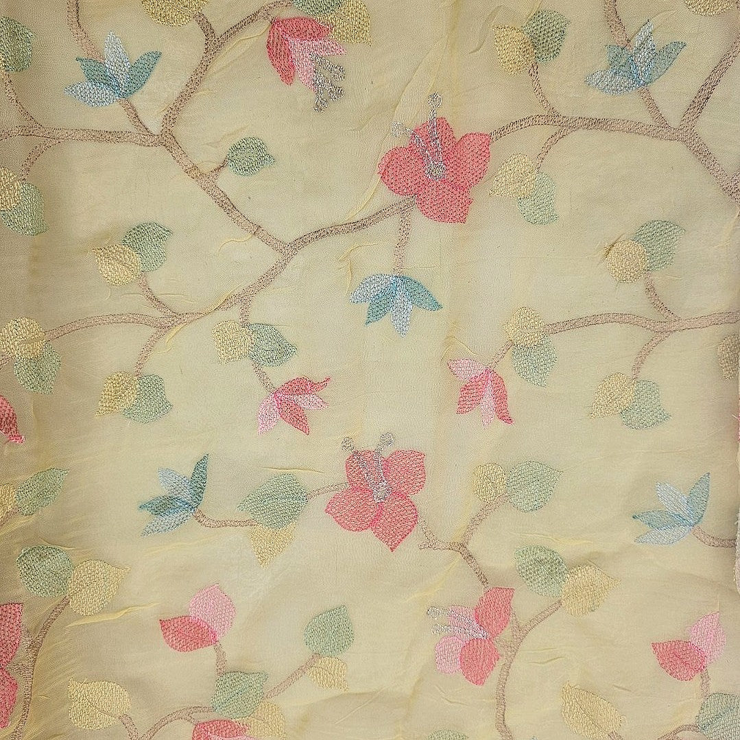 A close-up of a sheer cream-colored fabric featuring an embroidered floral vine pattern with pink blossoms, blue and green leaves, and brown branches.