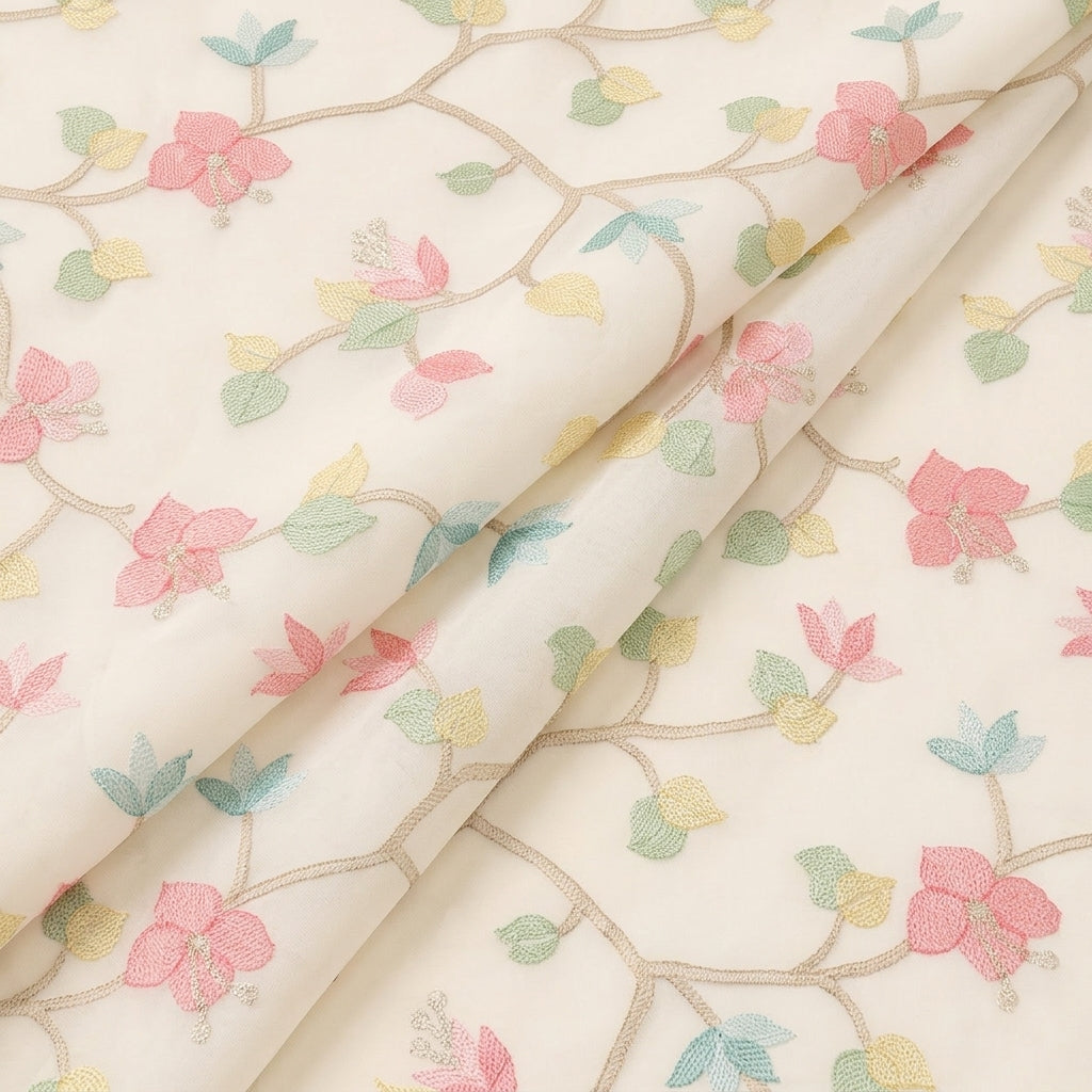 A close-up of white-colored fabric with a delicate embroidered floral pattern featuring pink blossoms, green and blue leaves, and beige branches.